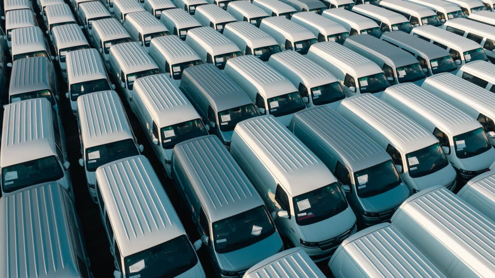 Aerial shot of multiple white vans parked in orderly rows in a lot under daylight.