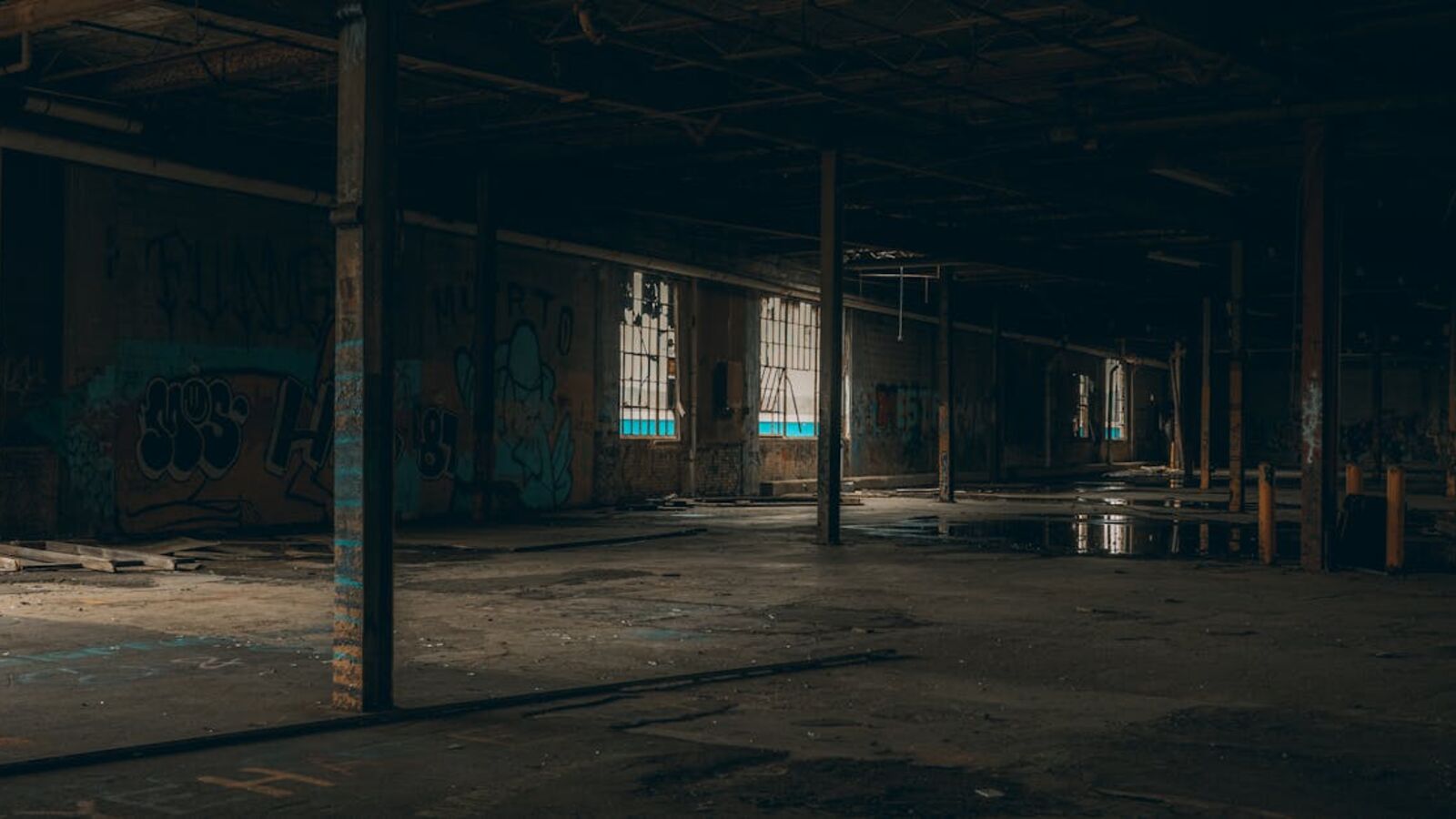 Dark and moody interior of a large abandoned warehouse with graffiti walls and broken windows.