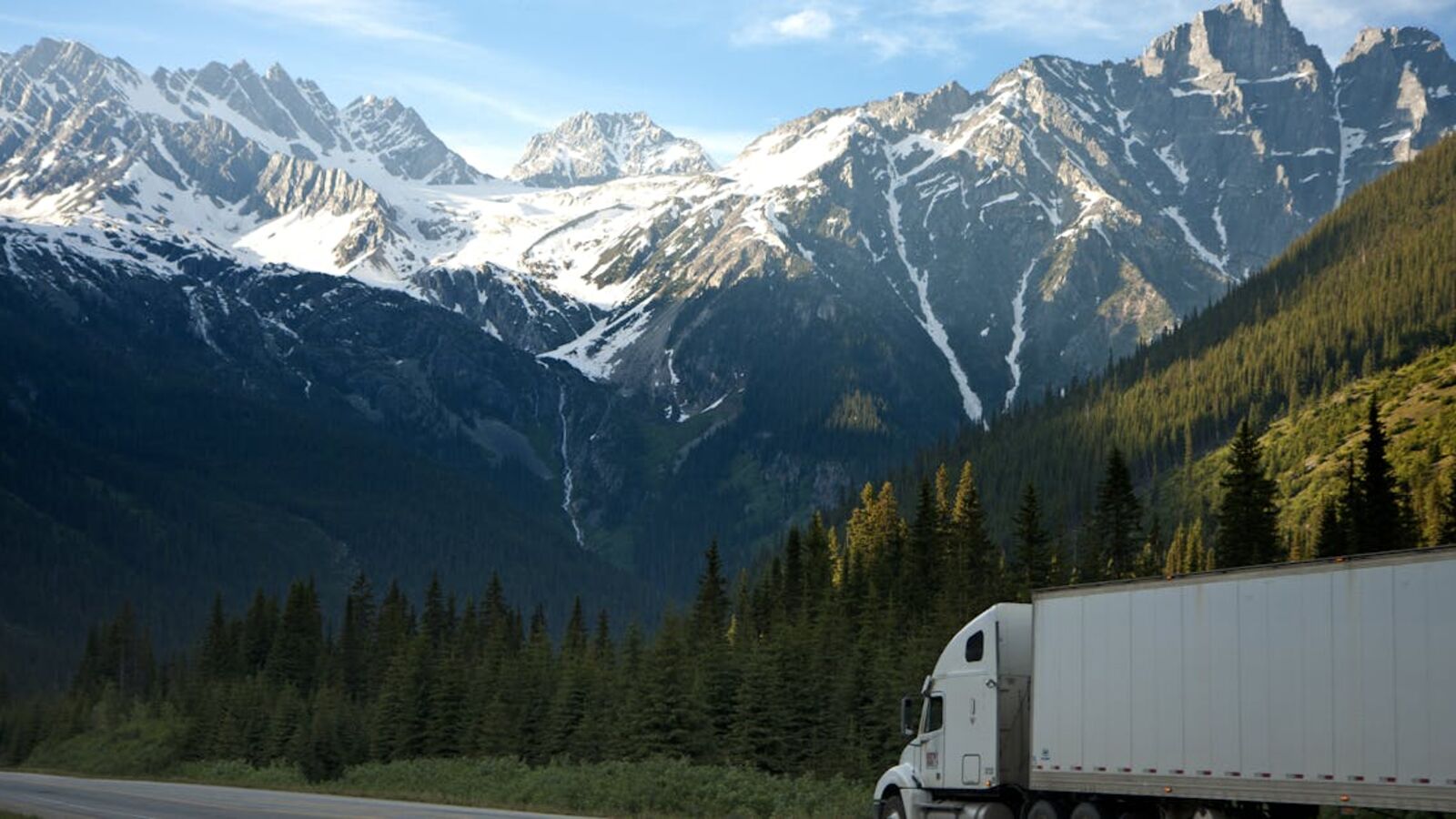 A semi-truck travels along a highway with snow-capped mountains in the background.