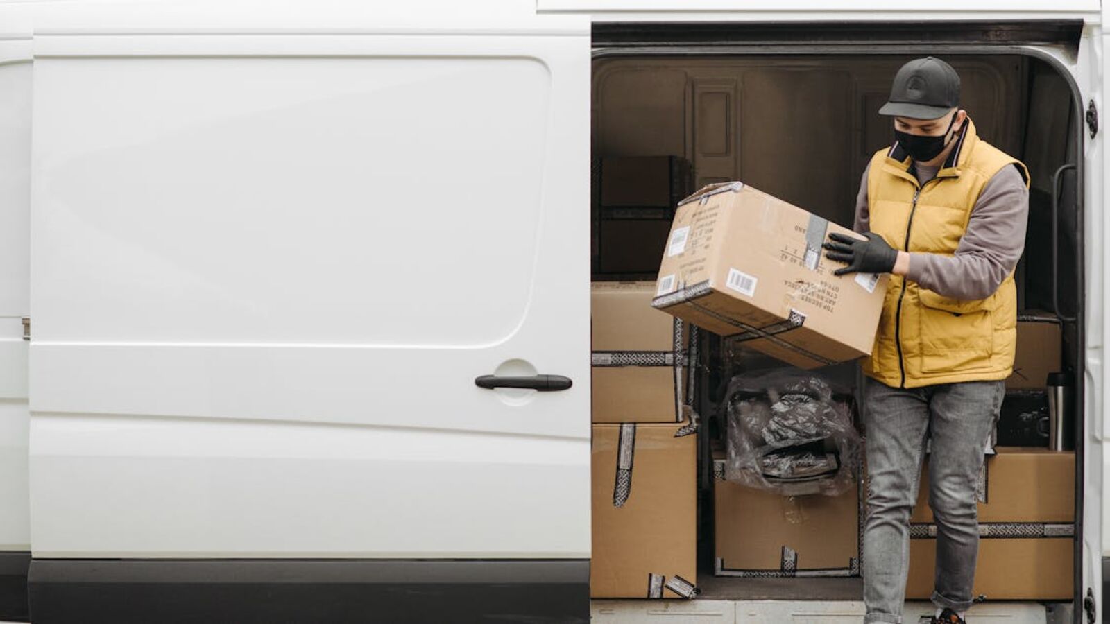Courier wearing mask unloads packages from white delivery van during the day.