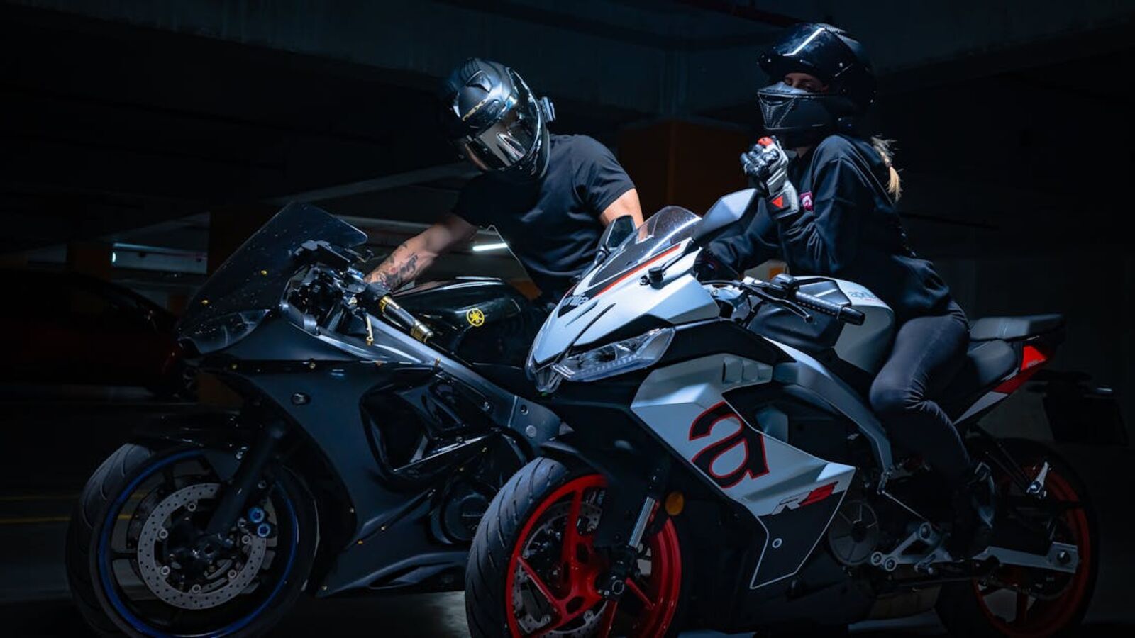 Two motorcyclists in helmets pose on sports bikes in a dimly lit garage.