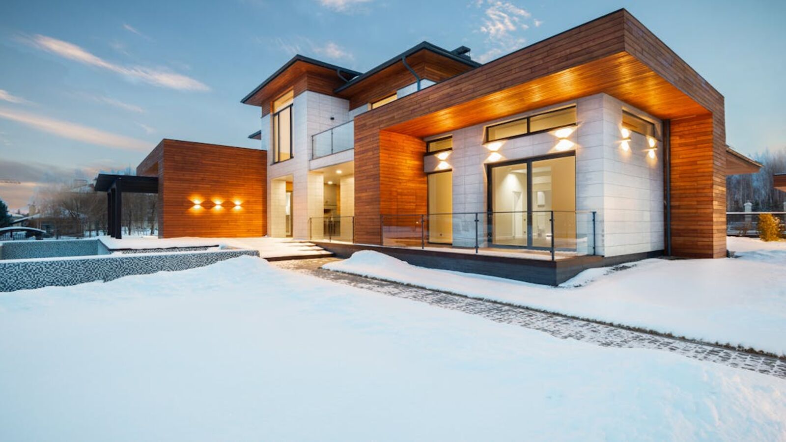 Exterior architecture of private suburban cottage house with stone and wooden facade and large windows overlooking spacious snow covered yard in winter day