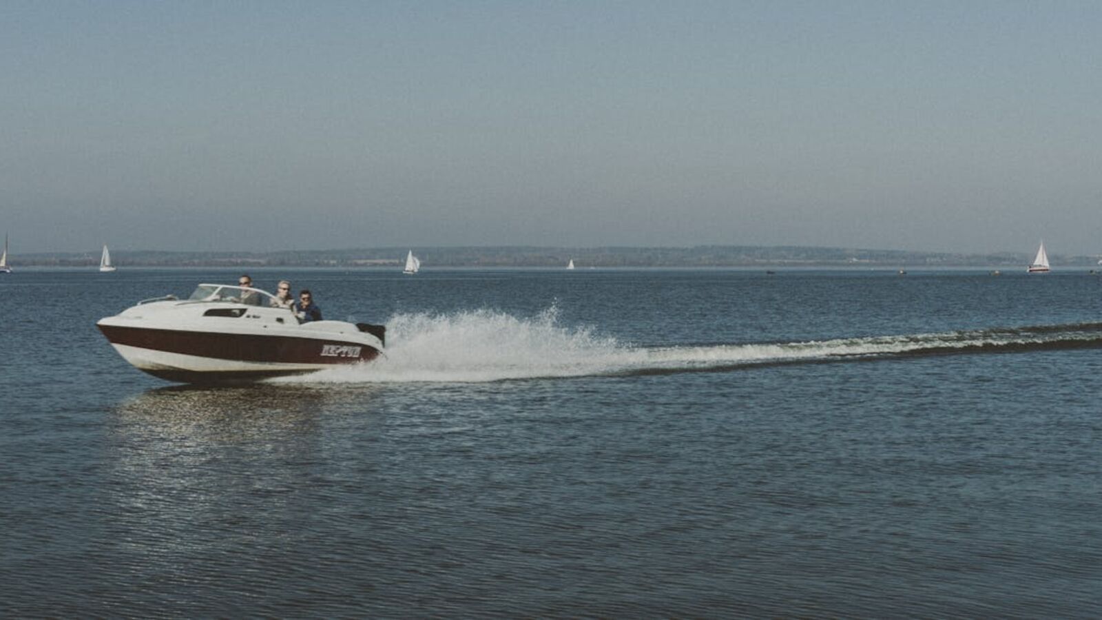 Motorboat speeding on a clear water body with sailboats in the distance on a sunny day.