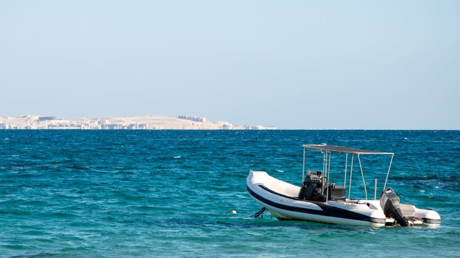 Inflatable RIB boat moored on clear blue sea with distant cityscape, under bright daylight.